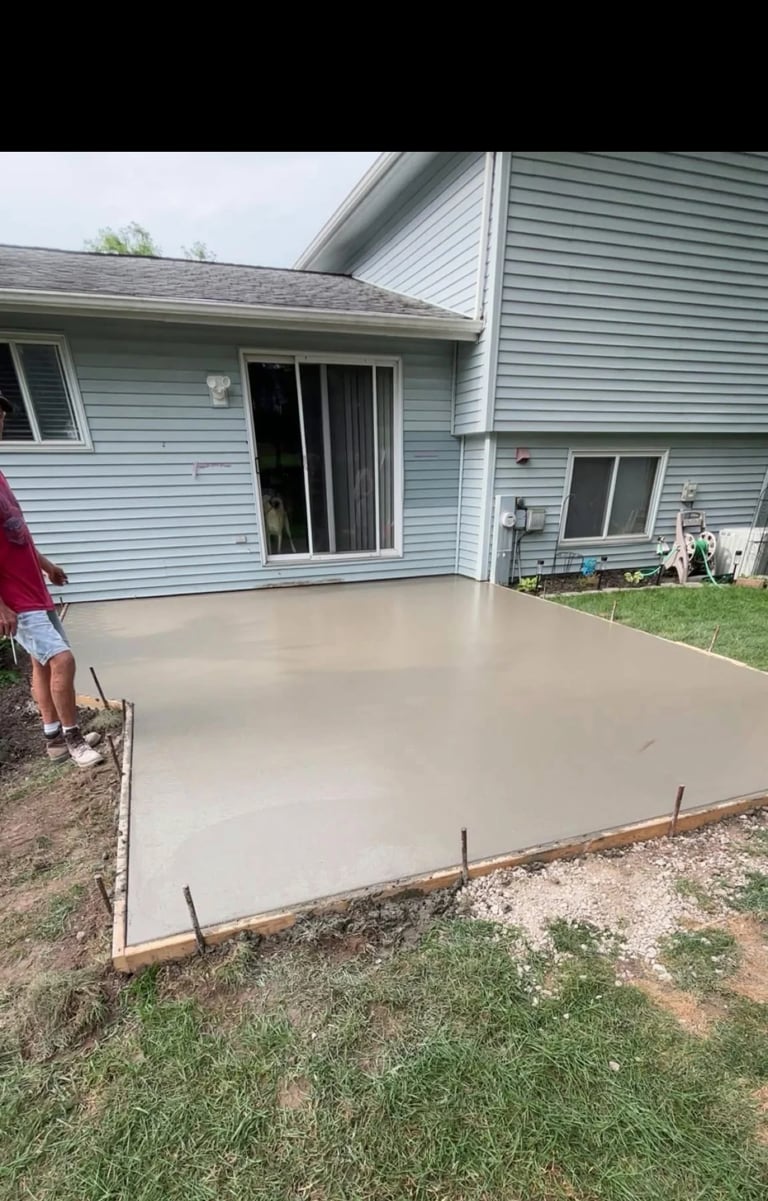 Newly poured concrete patio behind light blue house with person standing nearby and metal reinforcement rods visible