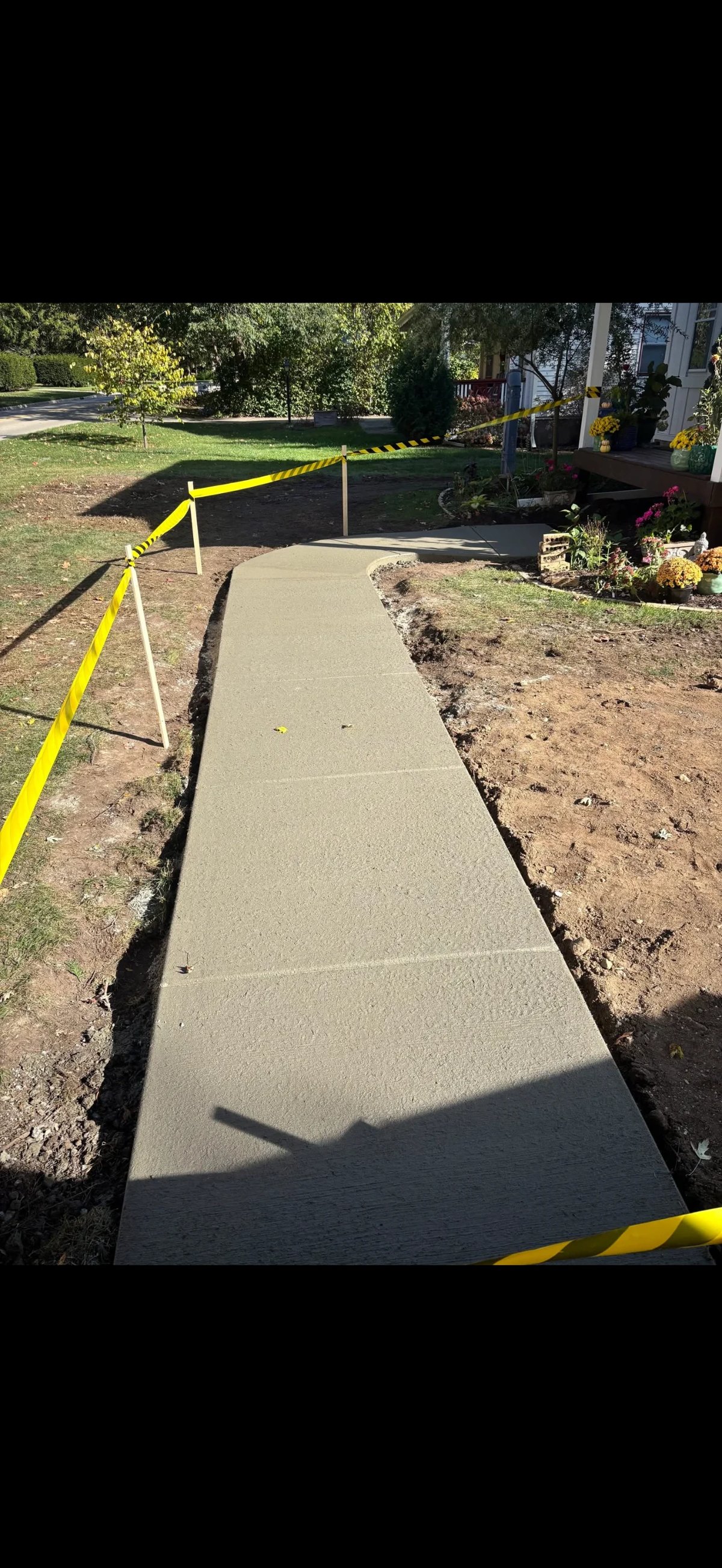 Newly poured concrete walkway cordoned off with yellow and black caution tape in a residential yard with landscaping