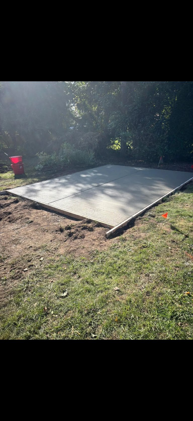 Concrete foundation pad laid on grass with dense tree line and construction markers in background