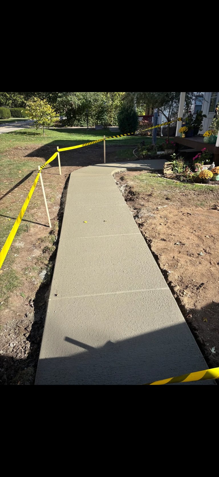 Newly poured concrete sidewalk bordered by yellow and black caution tape in a residential yard with freshly dug soil on sides